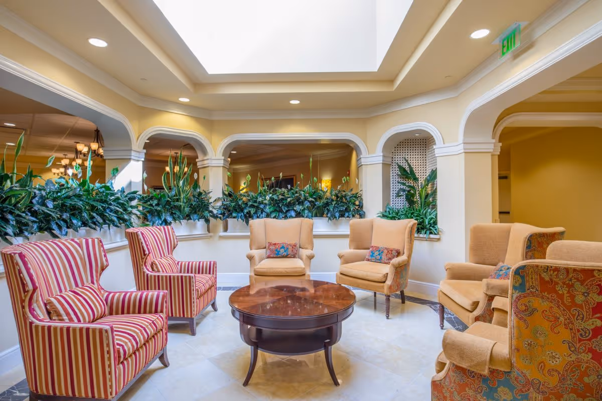 A cozy seating area in a senior living facility with six upholstered armchairs arranged around a round wooden coffee table. The chairs have different patterns, including striped and floral designs. The space features cream-colored walls, arched openings with plants on ledges, and a skylight above providing natural light.