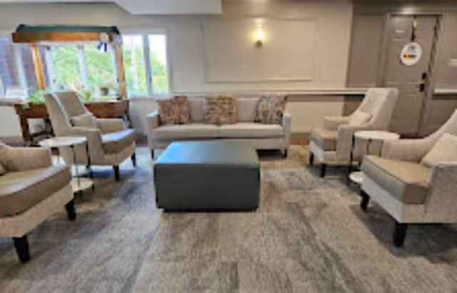 A cozy senior living common area with a beige sofa, four beige armchairs arranged around a large square ottoman, a window letting in natural light, and a shuffleboard table in the corner.