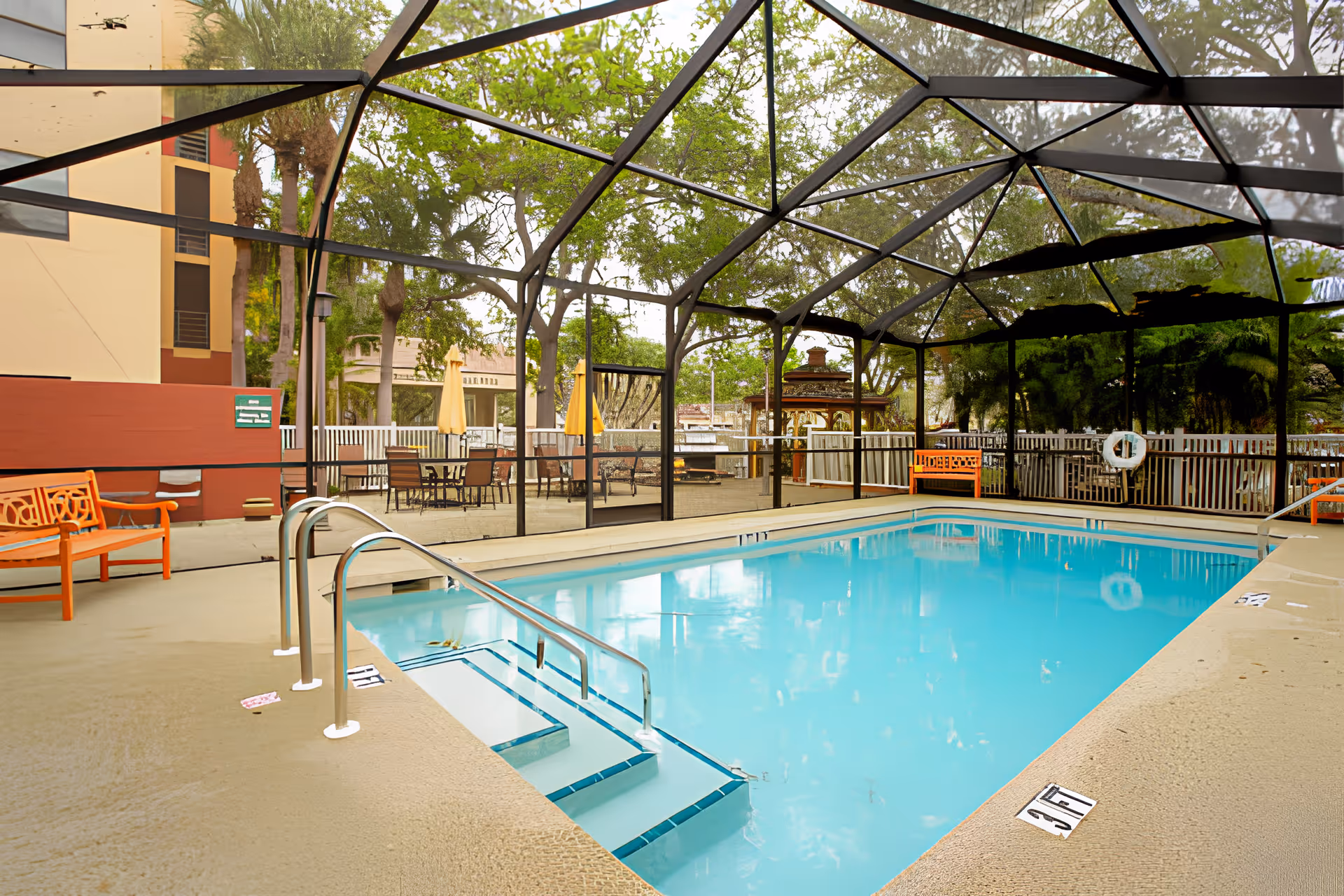 Enclosed outdoor swimming pool with steps and a metal handrail, surrounded by benches and patio seating under a screened pavilion.