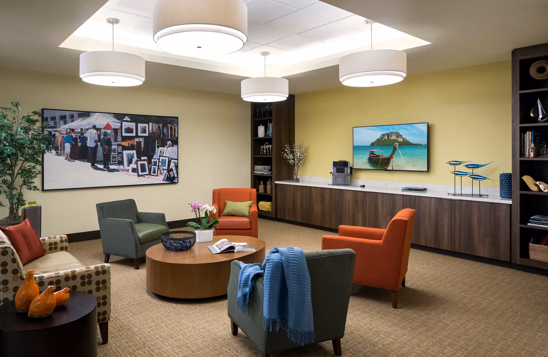A cozy living room area with a round wooden coffee table in the center, surrounded by four armchairs in various colors including orange, green, and patterned beige. The room has a beige carpet and light yellow walls. On one wall, there is a large framed photograph of people at an outdoor art market, and on the opposite wall, a flat-screen TV displays a scenic beach with a boat and island. The room is well-lit with three modern ceiling lights, and there are built-in wooden cabinets and shelves with decorative items and books.