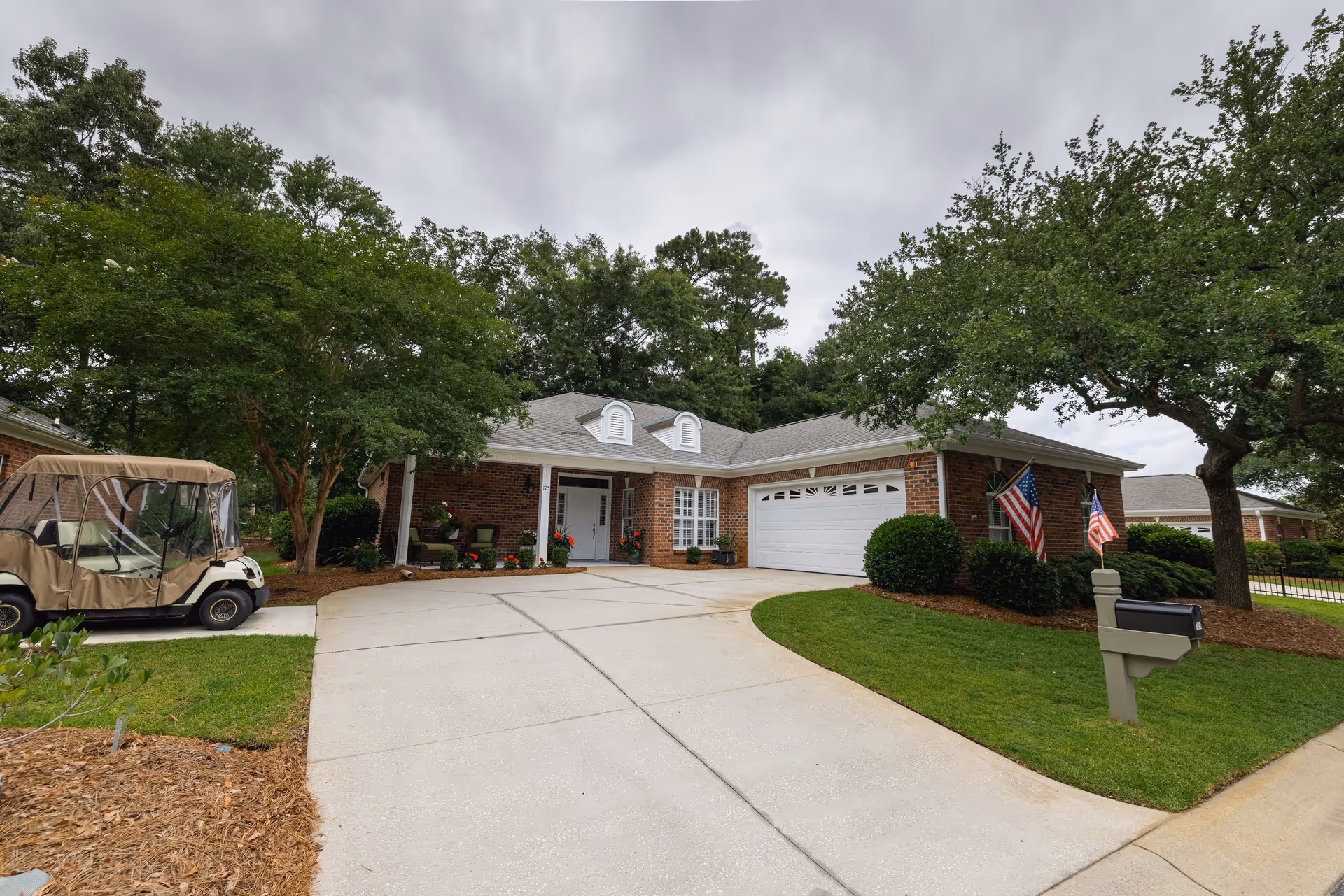 A single-story brick house with a white garage door and a white front door, surrounded by green trees and bushes. There is a concrete driveway leading up to the house, a golf cart parked on the left side, and two American flags displayed near the mailbox on the right. The sky is overcast.