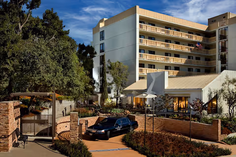Front entrance and facade of a multi-story senior living building with a gated driveway, landscaped courtyard and a parked black car under a blue sky.