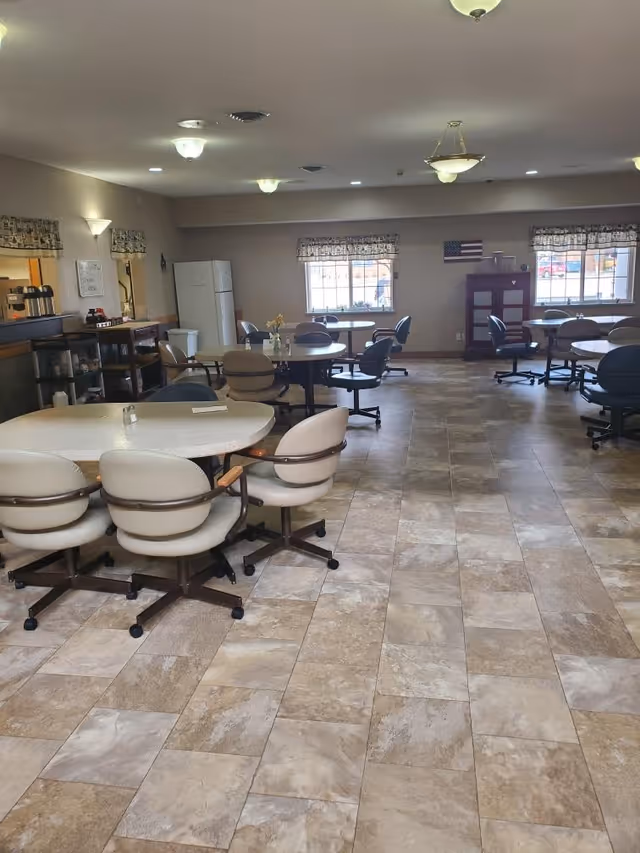 A spacious dining room with several round tables surrounded by cushioned swivel chairs on wheels. The room has tiled flooring, beige walls, and multiple ceiling lights. There are windows with patterned valances, a refrigerator, a coffee station, and a small cabinet with an American flag on the wall.