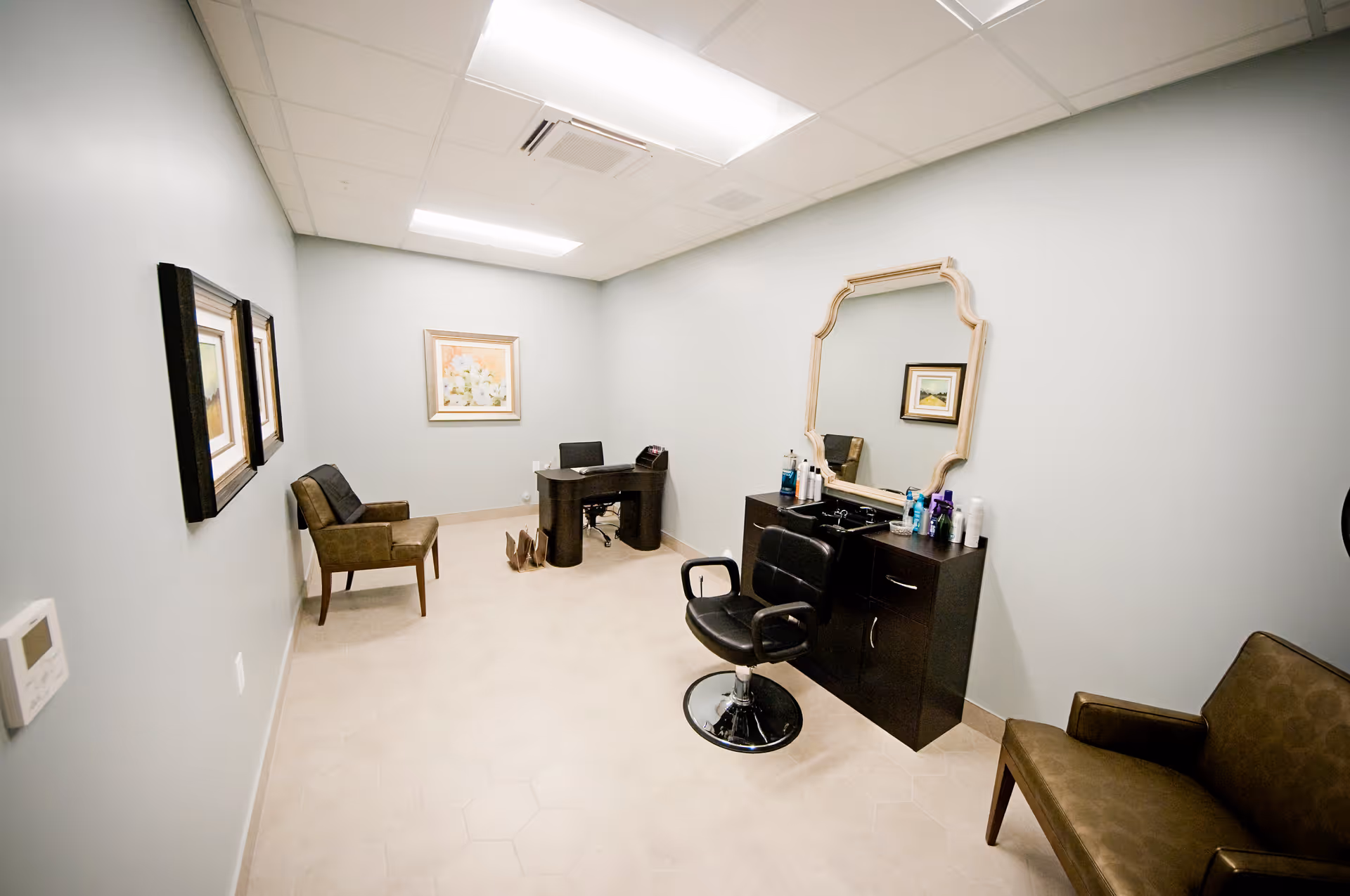 A small room with light-colored walls and tiled floor, featuring a black salon chair in front of a vanity with a large decorative mirror and various hair care products. There are two brown chairs along the walls and a small desk with a chair and a framed floral picture on the far wall.