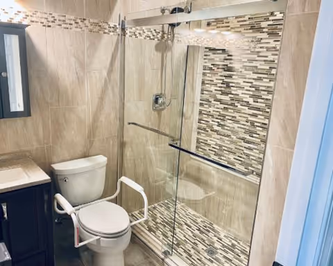 A modern bathroom featuring a toilet with white safety rails, a glass-enclosed shower with a tiled mosaic accent wall and floor, beige tiled walls, a dark wood vanity with a light-colored countertop, and a mirrored medicine cabinet.