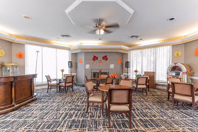 A bright and spacious common area with multiple round wooden tables and chairs arranged on a patterned carpet. The room features large windows with sheer curtains allowing natural light to fill the space. There is a fireplace decorated with autumn-themed decorations, including leaves and pumpkins. A ceiling fan is mounted in the center of the ceiling. On the left side, there is a wooden counter with a popcorn machine, and on the right side, a vintage jukebox is visible. Small flower arrangements are placed on each table.