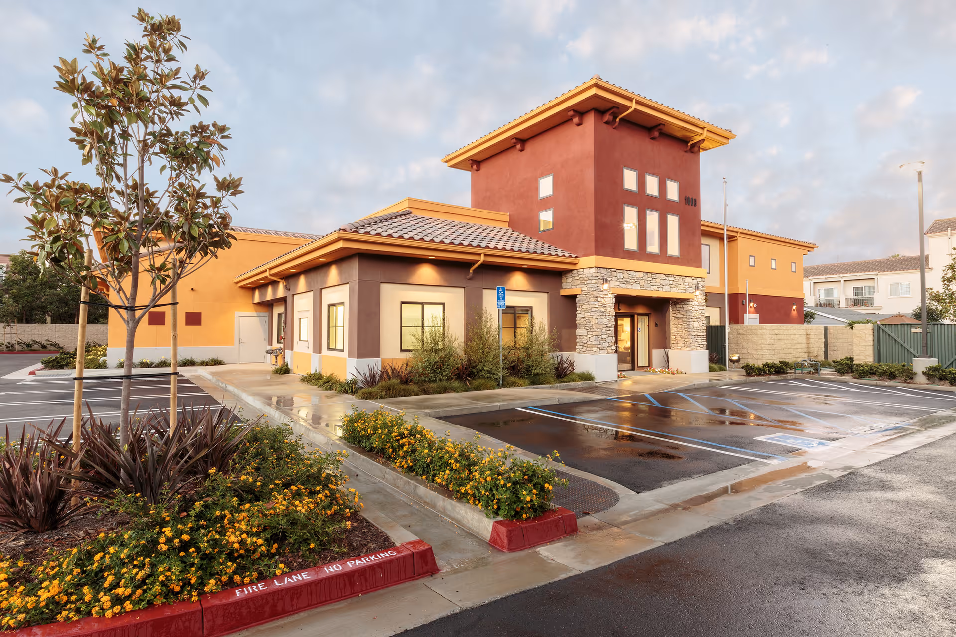 Exterior view of a modern senior living facility building with a mix of tan, brown, and red colors, stone accents around the entrance, and a tiled roof. The foreground shows a wet parking lot with marked handicapped spaces, landscaped areas with small trees and yellow flowers, and a cloudy sky above.