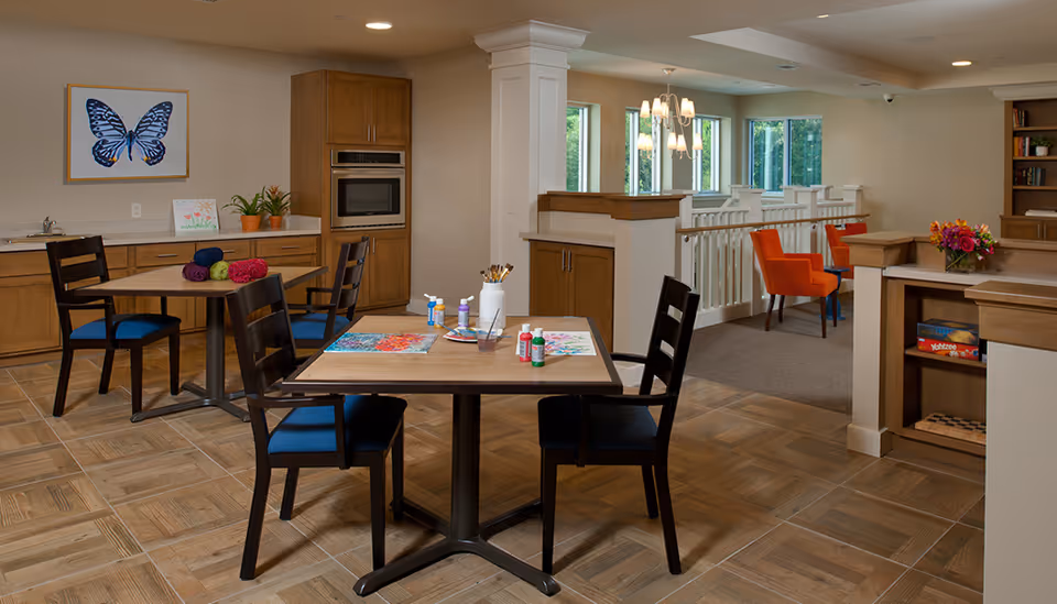 A bright, spacious activity room with wooden tables and chairs. One table has art supplies including paint bottles, brushes, and colorful paintings. Another table has yarn balls. The room features wooden cabinetry, a built-in microwave, a large butterfly painting on the wall, and a cozy seating area with orange chairs near windows letting in natural light.