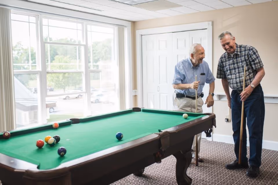 Two elderly men playing pool in a well-lit room with large windows. One man is wearing a light blue shirt and beige pants, while the other is in a plaid shirt and dark pants. They are both smiling and holding pool cues, standing next to a green pool table with several billiard balls on it.