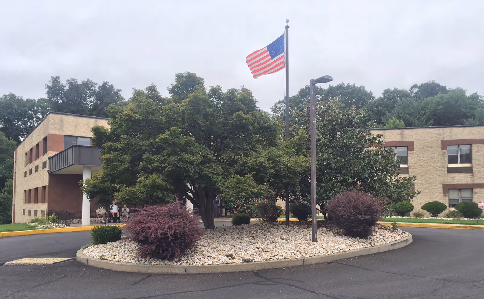 Entrance of a two-story brick health and rehabilitation facility with a circular driveway, landscaped island with trees and an American flag.