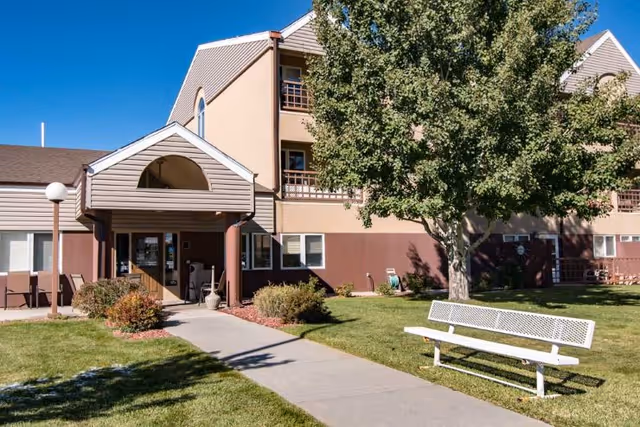 Exterior view of Edgewood Park Place, showing a multi-story building with beige and brown walls, a covered entrance with a walkway leading to it, a large tree, green lawn, bushes, and a white bench on the grass.