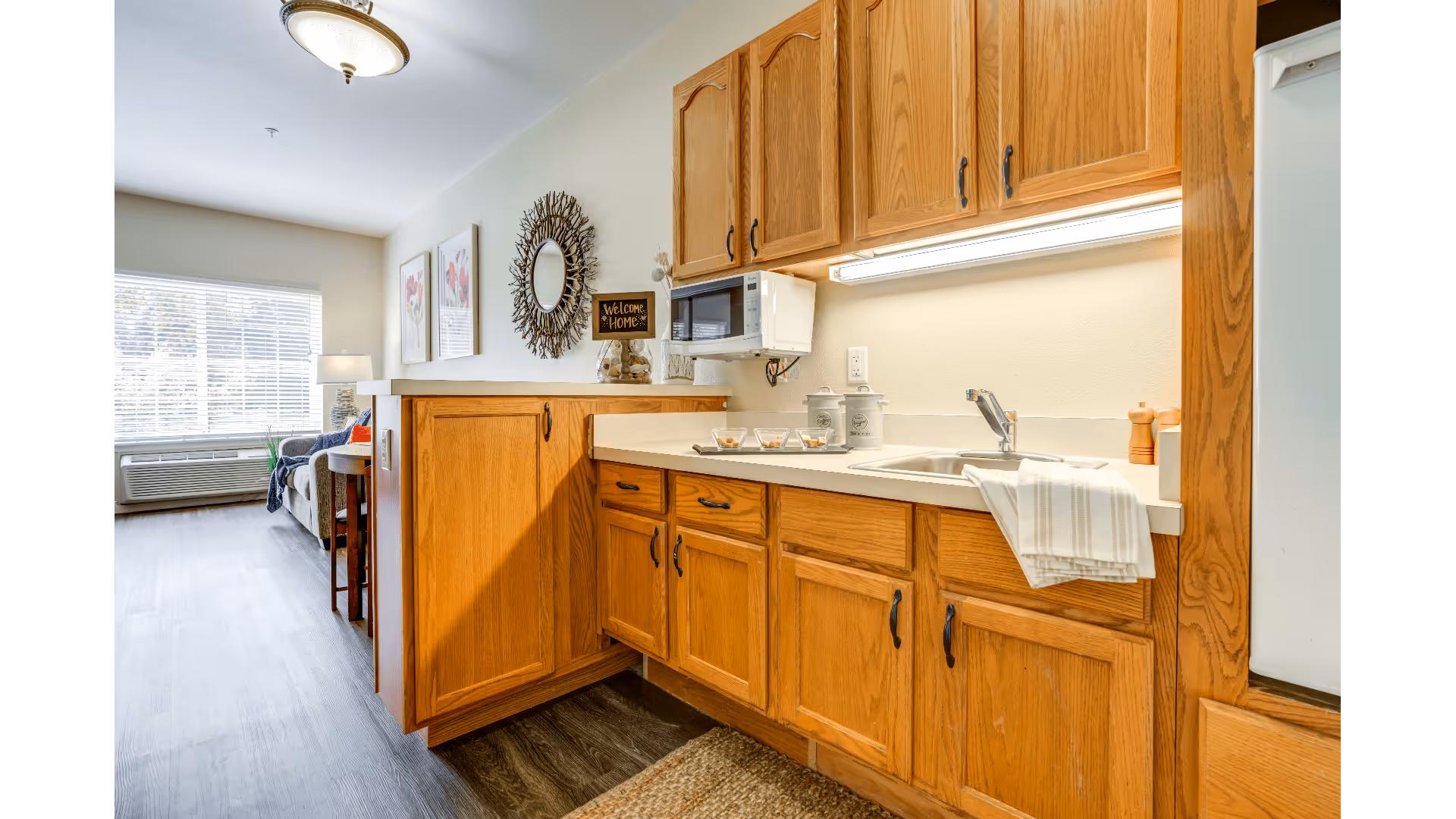A bright and clean kitchen area with wooden cabinets, a countertop with a sink, a microwave mounted under the upper cabinets, and a striped towel hanging over the sink. In the background, there is a living room area with a large window, a couch, a lamp, and wall decorations including framed floral artwork and a decorative round mirror with a 'Welcome Home' sign.