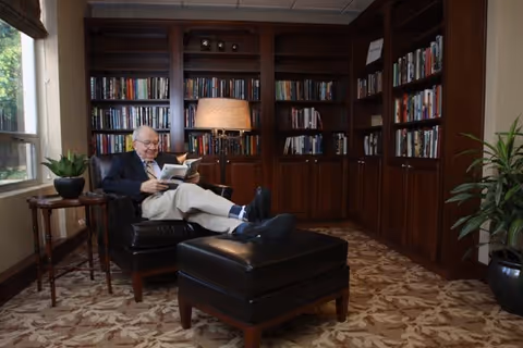 An elderly man sitting comfortably in a leather armchair with his feet resting on a matching ottoman, reading a book in a cozy room with wooden bookshelves filled with books, a table lamp, a small side table with a plant, and a large potted plant near the window.