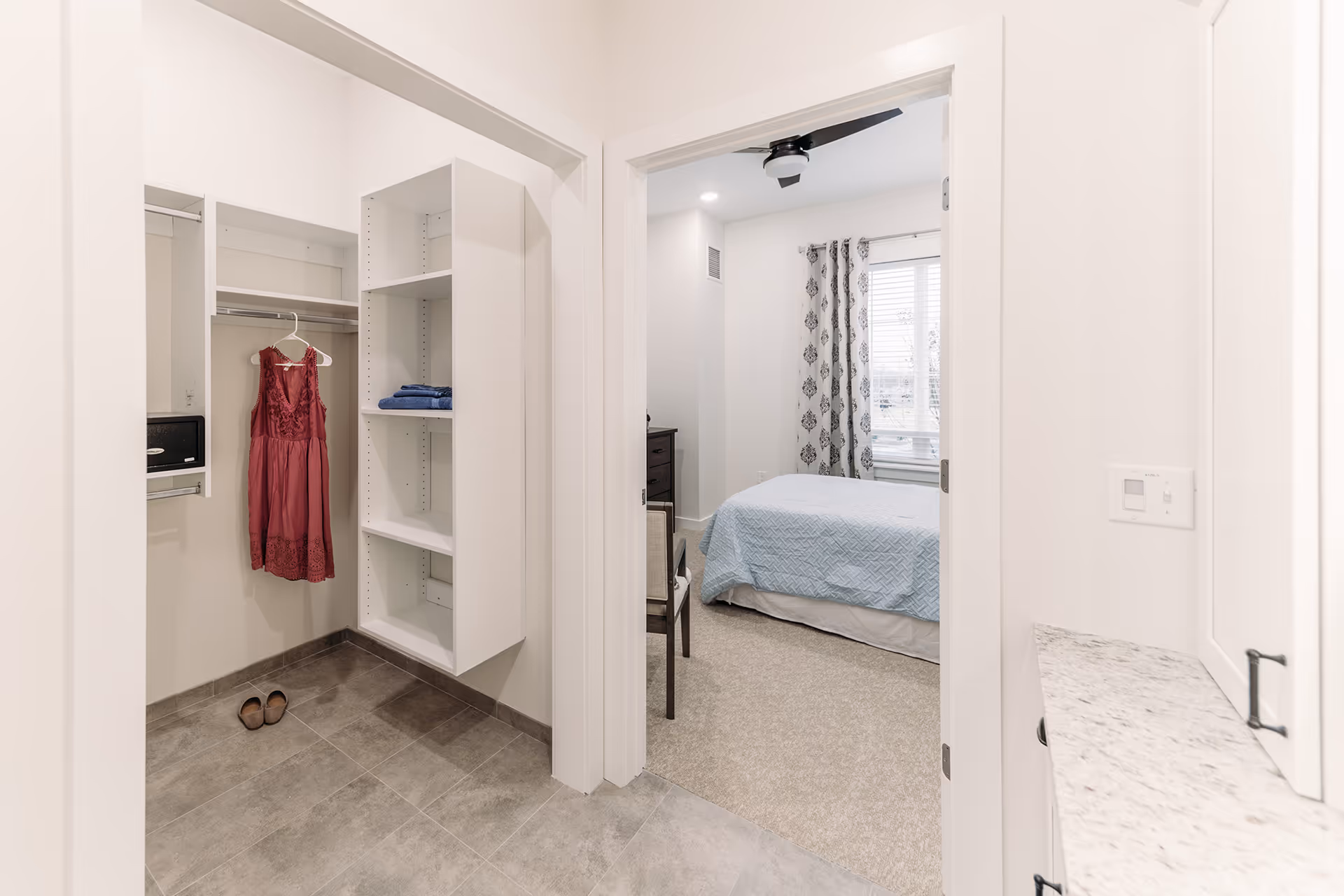 View of a walk-in closet with a red dress hanging and shelves with folded towels, adjacent to a bedroom with a bed covered in a light blue quilt, a chair, a window with patterned curtains, and a ceiling fan.