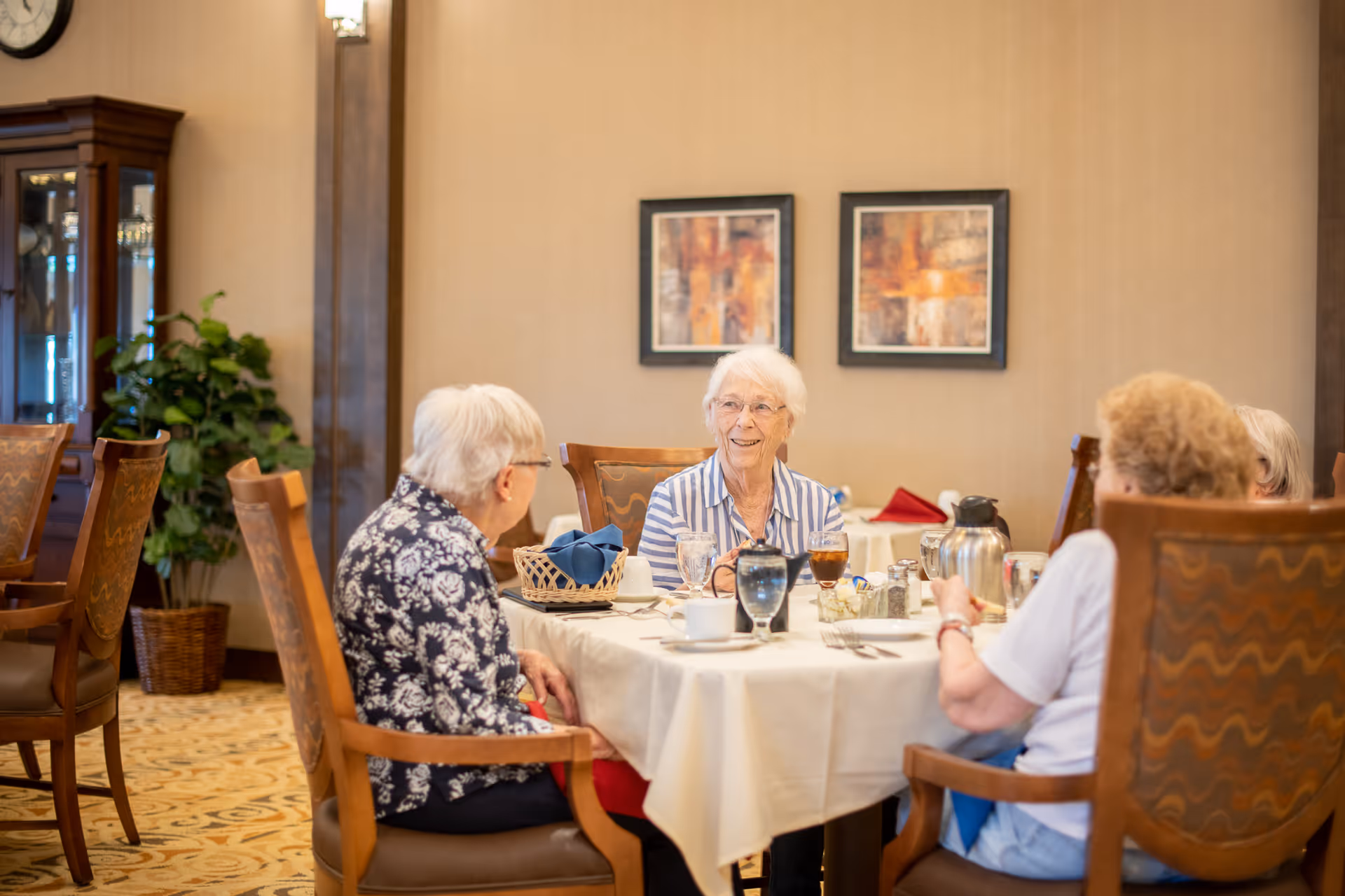 Four elderly women sitting around a dining table in a well-lit room, engaged in conversation. The table is set with cups, glasses, and a coffee pot. The room has wooden chairs, a patterned carpet, framed artwork on the wall, and a plant in the corner.