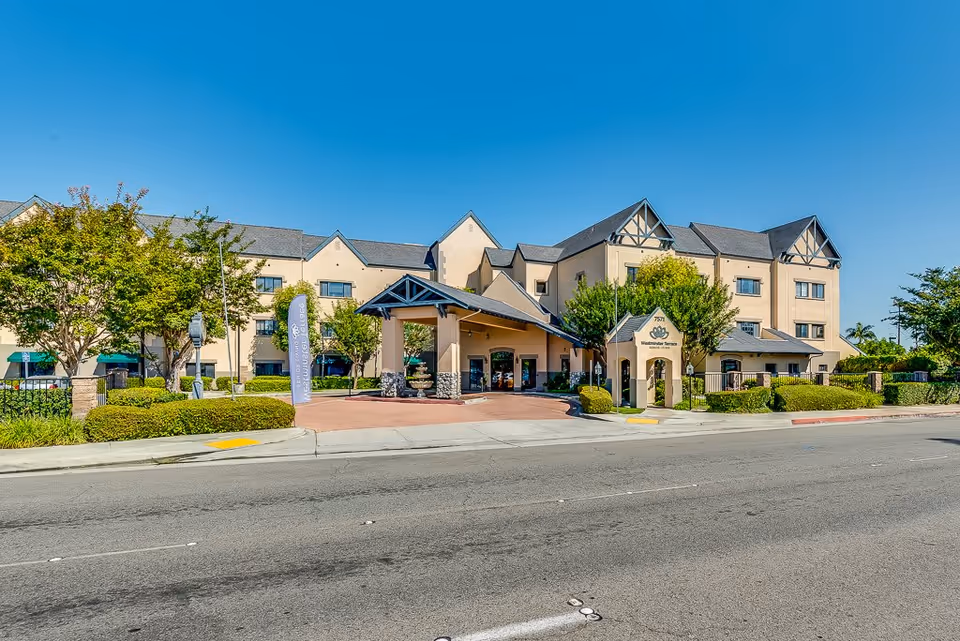 Exterior view of Westminster Terrace Senior Living facility showing a three-story beige building with a covered entrance, trees, bushes, and a clear blue sky.
