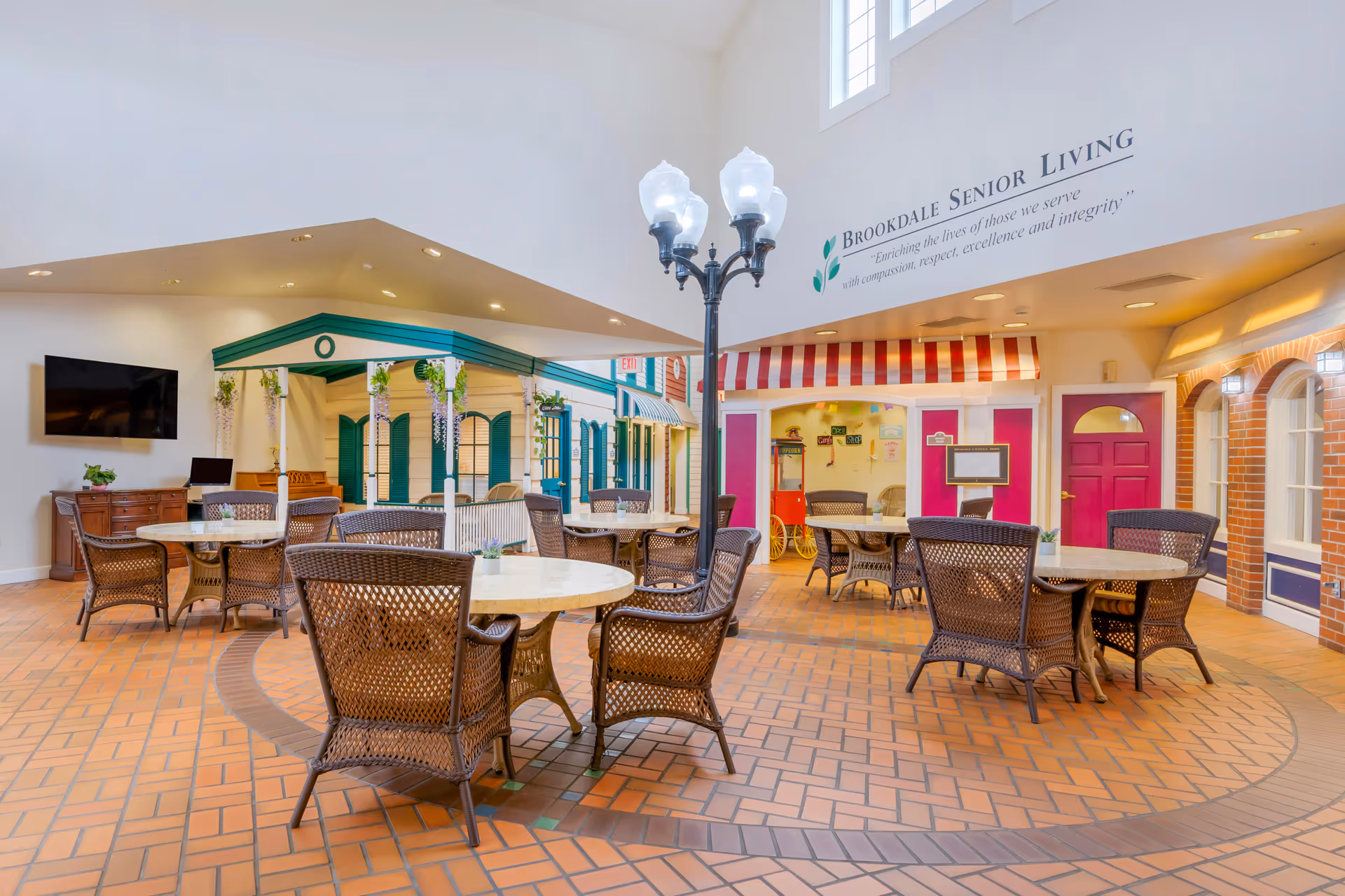 Indoor seating area with round tables and wicker chairs arranged on a tiled floor. The space features a decorative streetlamp in the center and storefront-style facades with colorful doors and awnings. A sign on the wall reads 'Brookdale Senior Living' with a mission statement underneath.