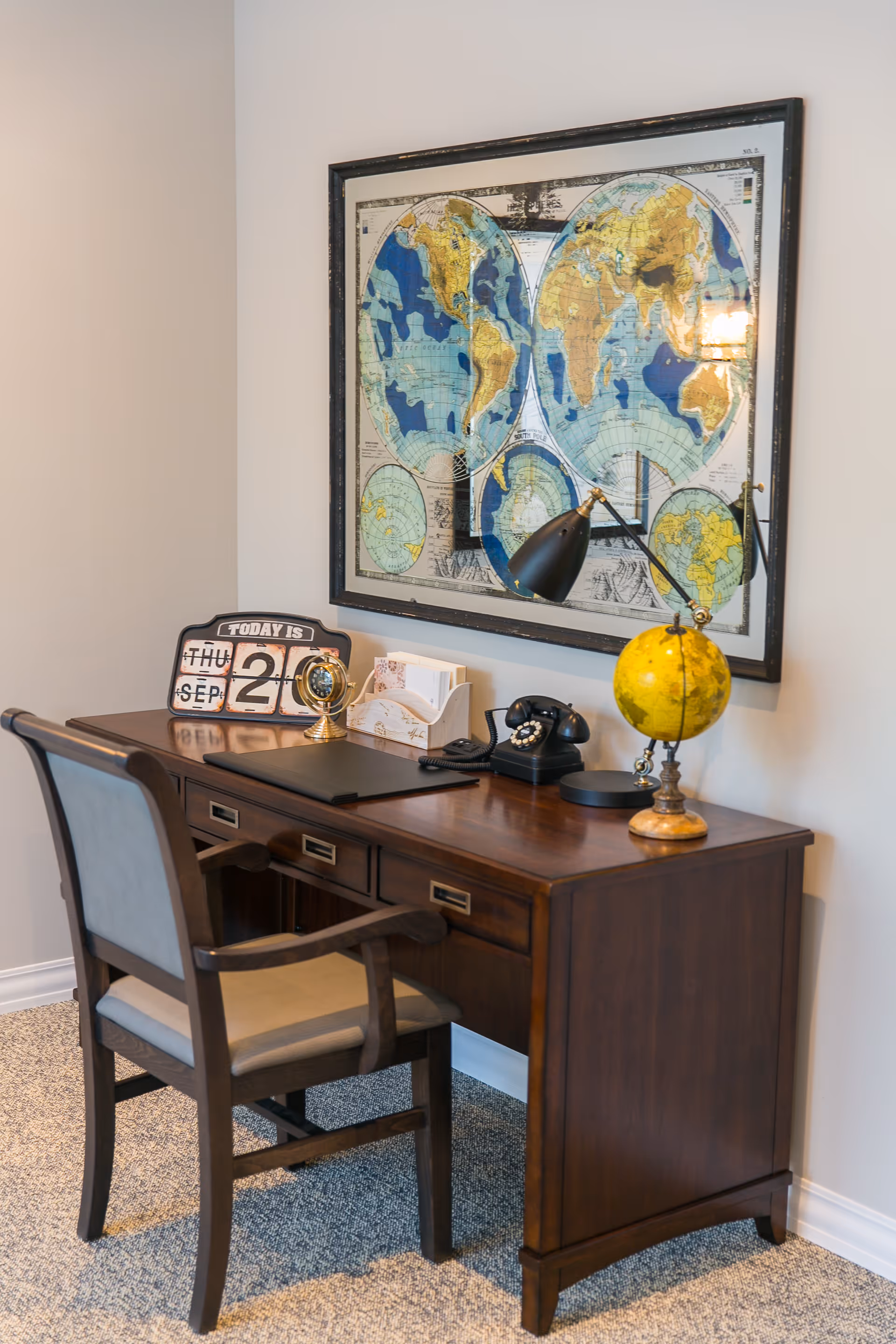 A wooden desk with a matching chair in a room. On the desk are a black desk lamp, a vintage rotary phone, a small yellow globe, a calendar showing Thursday, September 20, a small clock, and a letter holder. Above the desk is a large framed world map with multiple hemispheres.