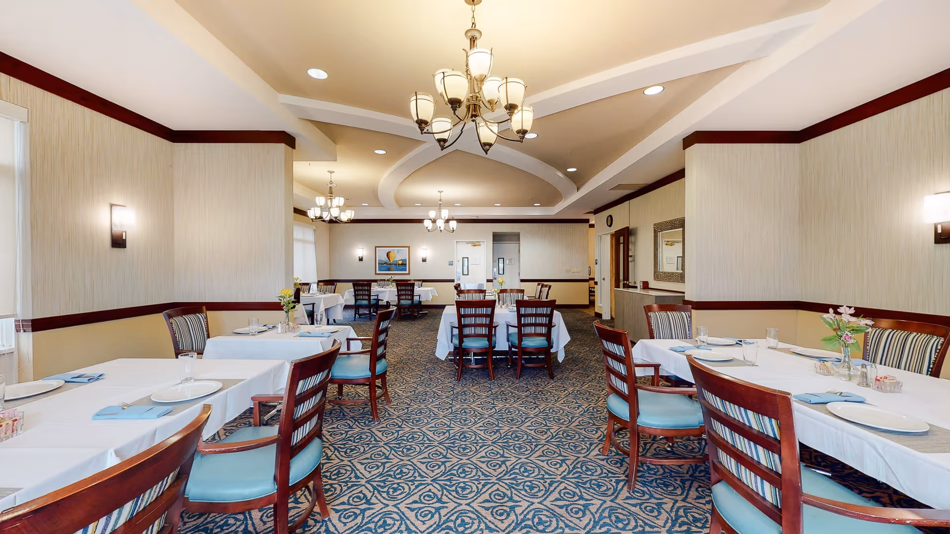 A spacious dining room with multiple tables covered in white tablecloths, each set with plates, glasses, and napkins. The room features wooden chairs with blue cushions, patterned carpet, and elegant chandeliers hanging from the ceiling. The walls are light-colored with dark wood trim, and there is a painting and a mirror on the walls.
