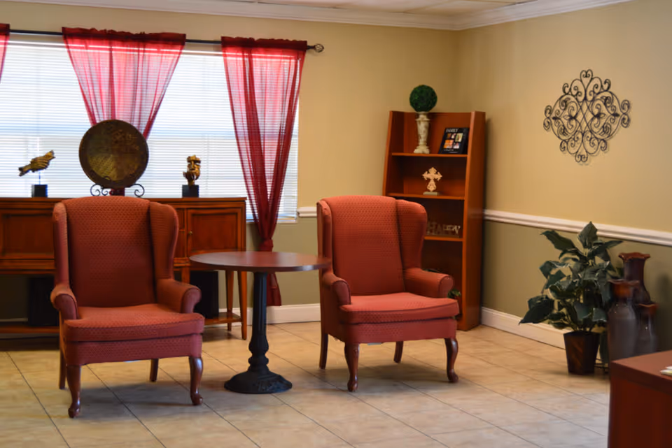 A cozy sitting area with two red upholstered armchairs positioned around a small round wooden table. Behind the chairs is a wooden sideboard with decorative items including a large round plate and small sculptures. Red sheer curtains hang over a window with blinds. To the right, there is a wooden bookshelf with decorative items and a green potted plant next to two large vases. The walls are painted beige with white trim and a decorative metal wall hanging is visible.