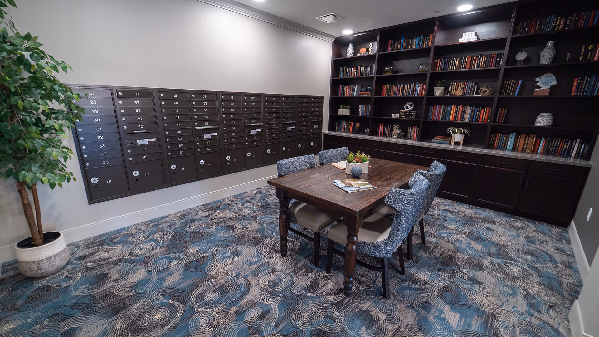 Interior common area with a wooden table and chairs, a wall of mailboxes on the left and built-in bookshelves on the right.