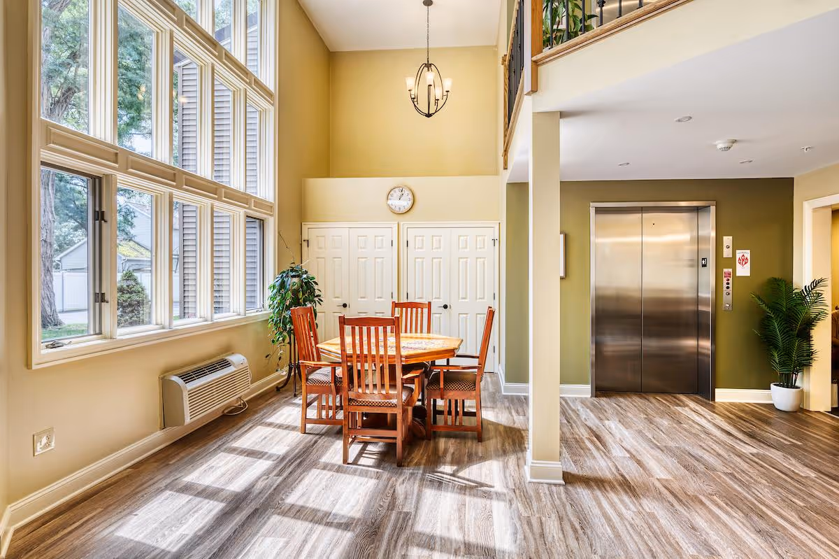A bright interior space with large windows letting in natural light, featuring a wooden table with six chairs in the center. There is a clock on the wall above two white doors, a green potted plant near the windows, and an elevator with closed stainless steel doors on the right side. The floor is wood-patterned, and the walls are painted in light beige and green tones.