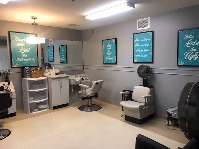 Interior view of a salon area in a senior living facility with salon chairs, a hair washing station, and framed inspirational quotes on the walls. The room has light gray walls, a beige floor, and a modern light fixture.