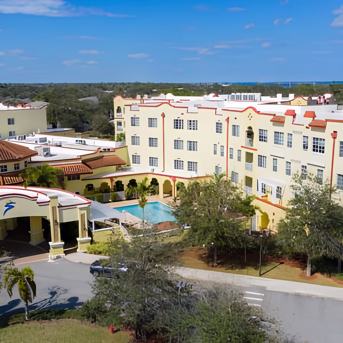 Exterior view of Discovery Village At Melbourne, showing a multi-story building with cream-colored walls and red trim. The building surrounds a swimming pool area with trees and greenery around. The sky is clear and blue.