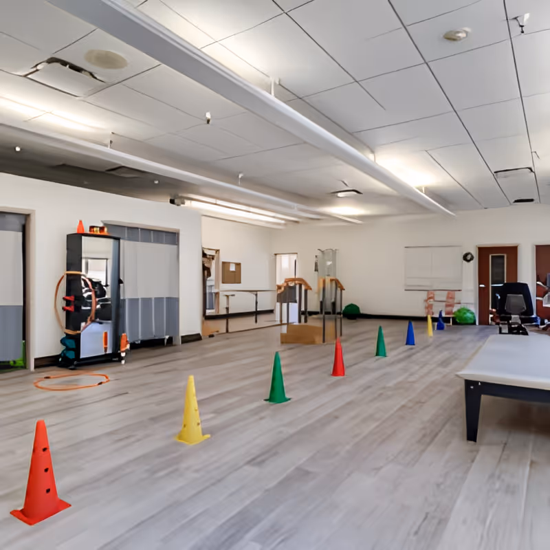 A spacious room with light wood flooring and white walls, featuring a row of colorful cones arranged in a line on the floor. The room has large mirrors on one wall, exercise equipment, and a bench. The ceiling has fluorescent lighting and acoustic tiles.