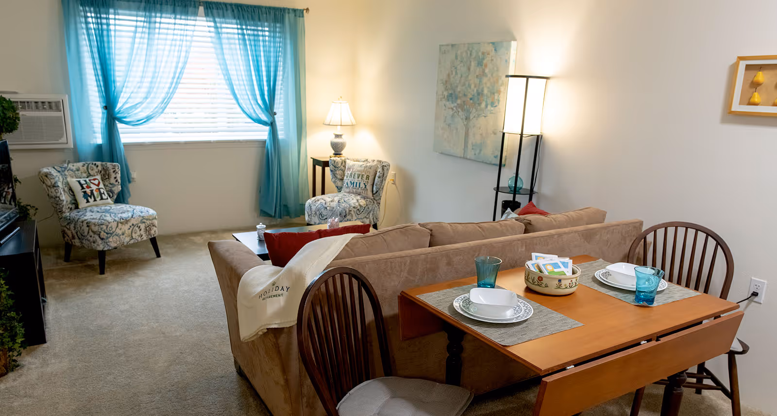 A cozy living room and dining area with a beige couch, two patterned armchairs with decorative pillows, a wooden dining table set for two with plates and glasses, a floor lamp, a table lamp on a side table, blue curtains on a window, and wall art.