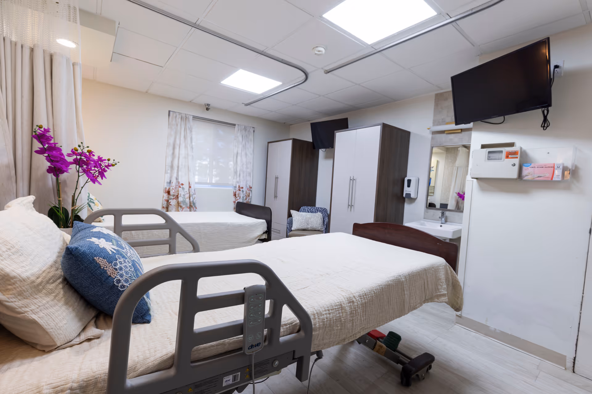 A nursing facility room with two hospital beds, each made with white bedding and pillows. There is a purple orchid plant on a small table next to one bed. The room has two wardrobes, a wall-mounted TV, a sink with a mirror, and a window with floral curtains. The floor is light-colored wood, and the ceiling has recessed lighting.