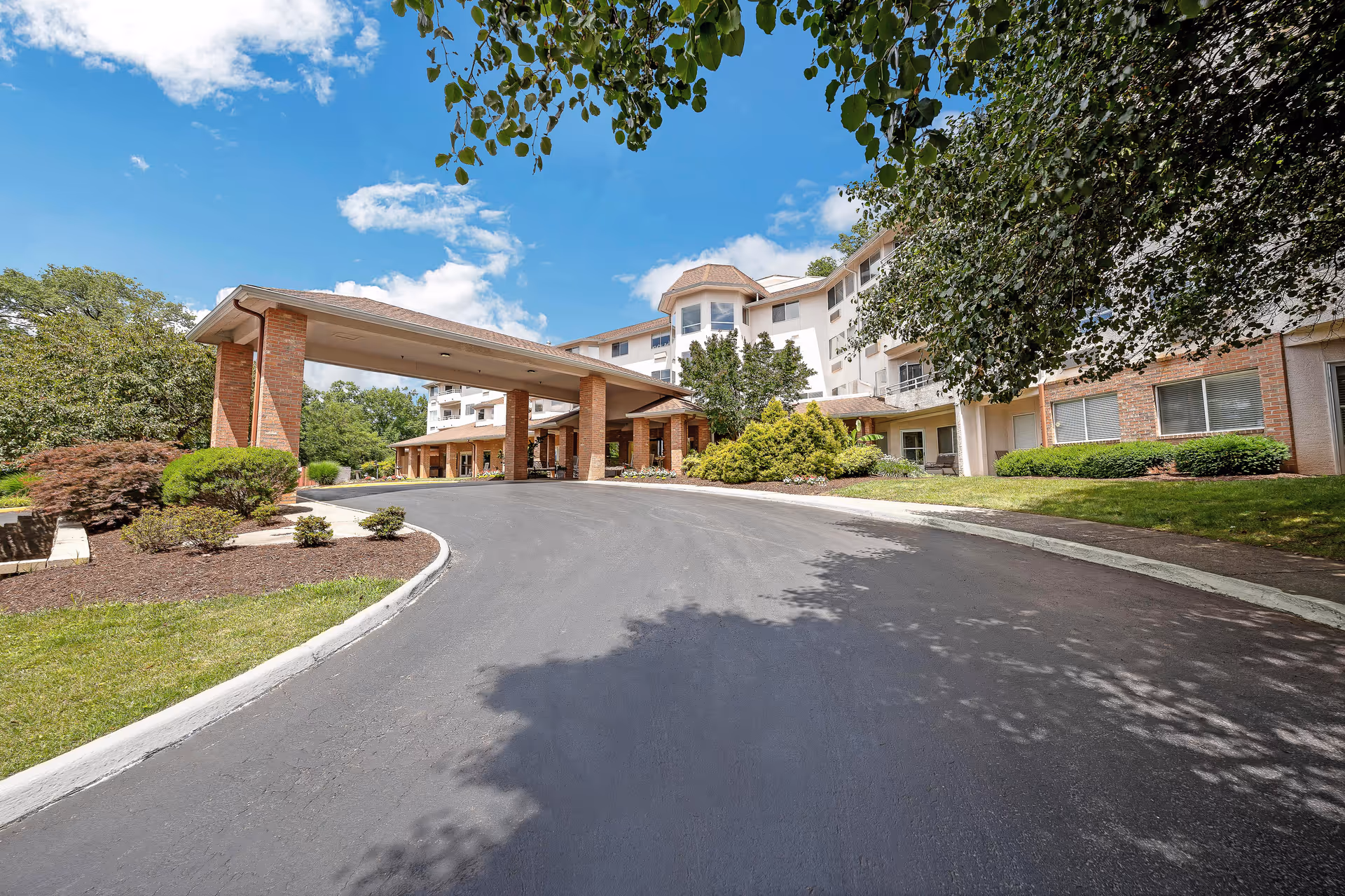 Exterior view of Holiday Elm Park Estates senior living facility showing a curved driveway leading to a covered entrance with brick pillars, surrounded by landscaped greenery and trees under a blue sky with some clouds.