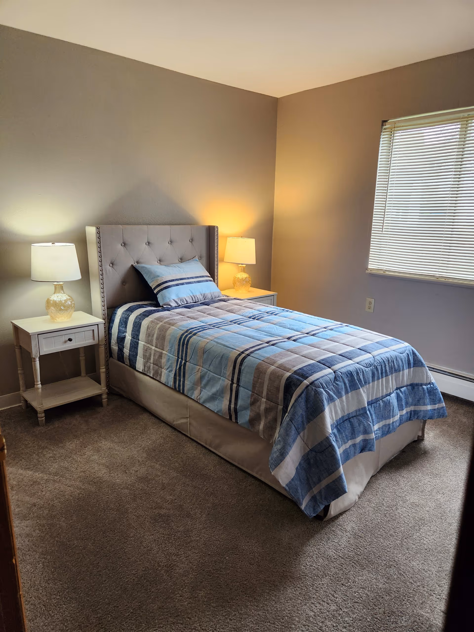 A neatly made single bed with a blue, gray, and white striped comforter and matching pillow in a bedroom. The bed has a tufted headboard and is flanked by two white nightstands, each with a lamp. There is a window with closed blinds on the right wall, and the room has beige walls and carpeted floor.