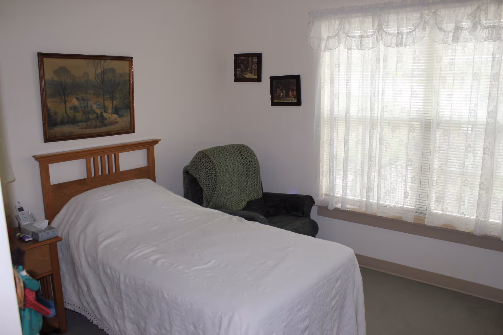 A small bedroom with a single bed covered in a white quilt. Next to the bed is a wooden nightstand with a telephone and a box of tissues. A green armchair with a knitted green blanket draped over it is positioned near a window with white lace curtains. The walls are decorated with three framed pictures.