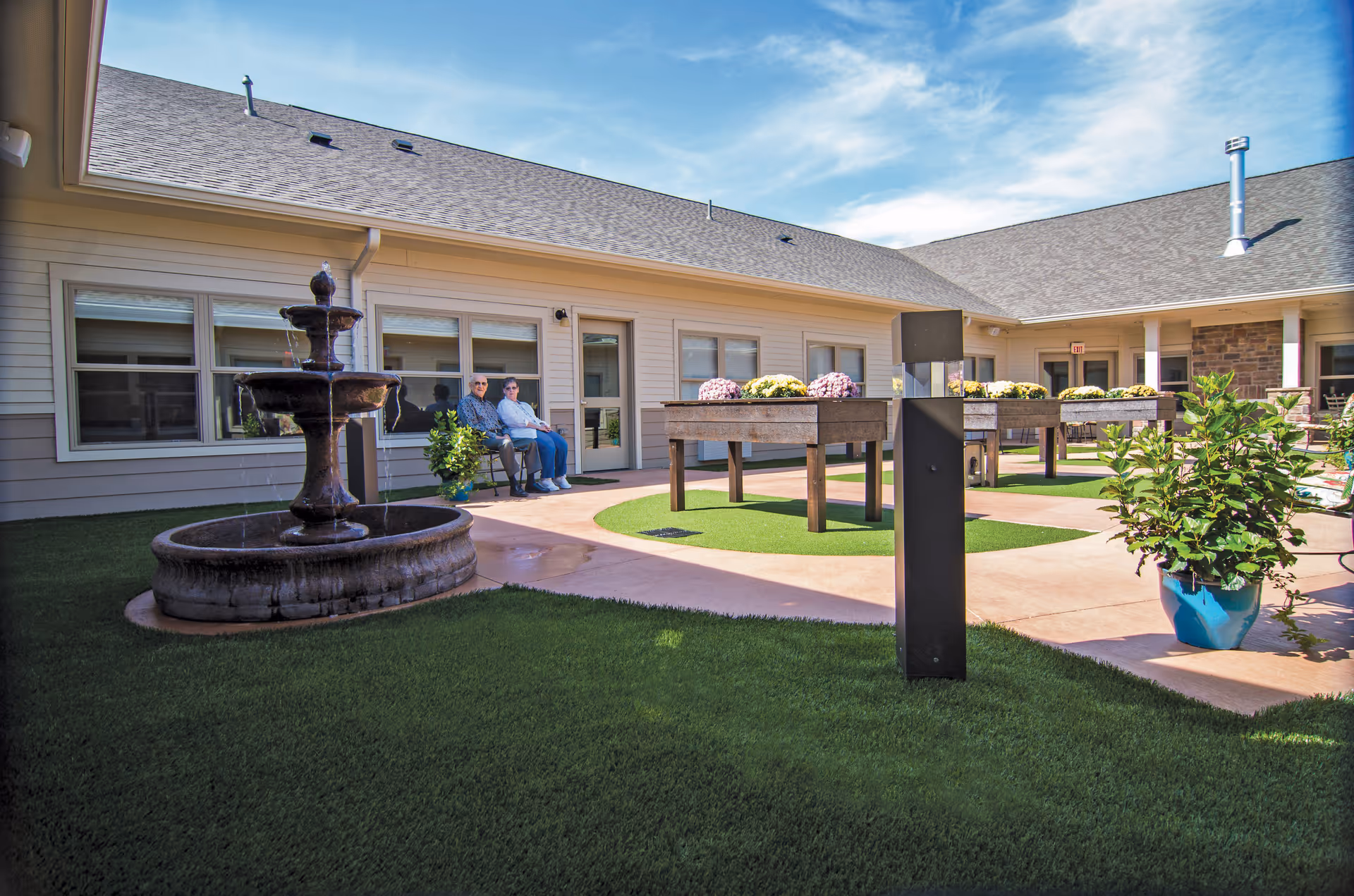 Outdoor courtyard area at Primrose Memory Care of Anderson featuring a multi-tiered water fountain, two people sitting on a bench near the building, raised garden beds with flowers, potted plants, and a clear blue sky.