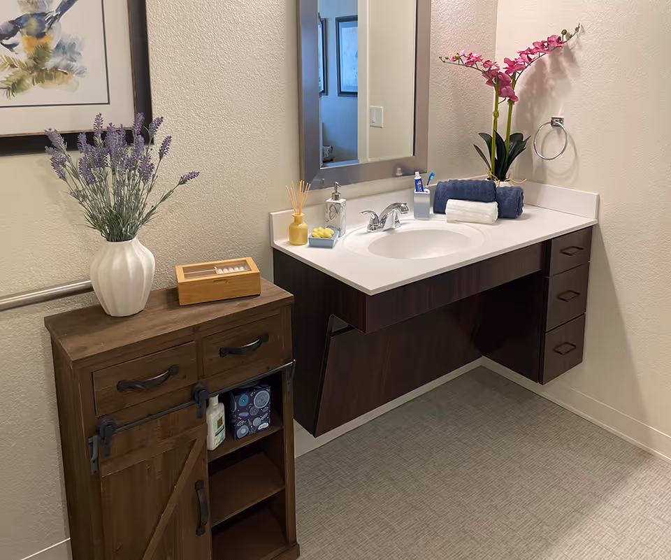 A clean and modern bathroom vanity area with a white countertop and dark wood cabinetry. On the countertop are a soap dispenser, a toothbrush holder with toothbrushes, a small tray with yellow soap, a rolled white towel, a rolled dark blue towel, and a potted orchid with pink flowers. To the left of the vanity is a small wooden cabinet with two drawers and a sliding barn-style door, topped with a white vase holding lavender flowers and a wooden box. A large mirror is mounted on the wall above the sink, and a towel ring is attached to the wall on the right side.