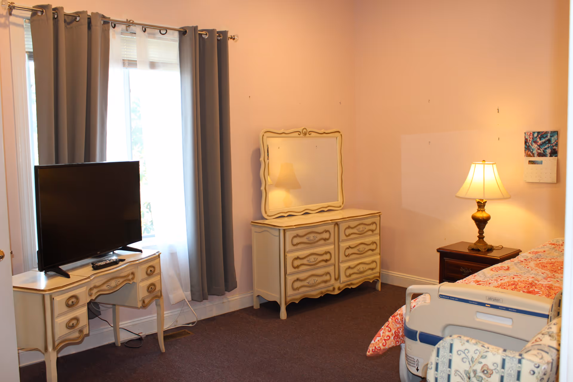 A cozy bedroom with a TV on a small table, a vintage dresser with mirror, a bedside lamp, and a bed with patterned bedding.