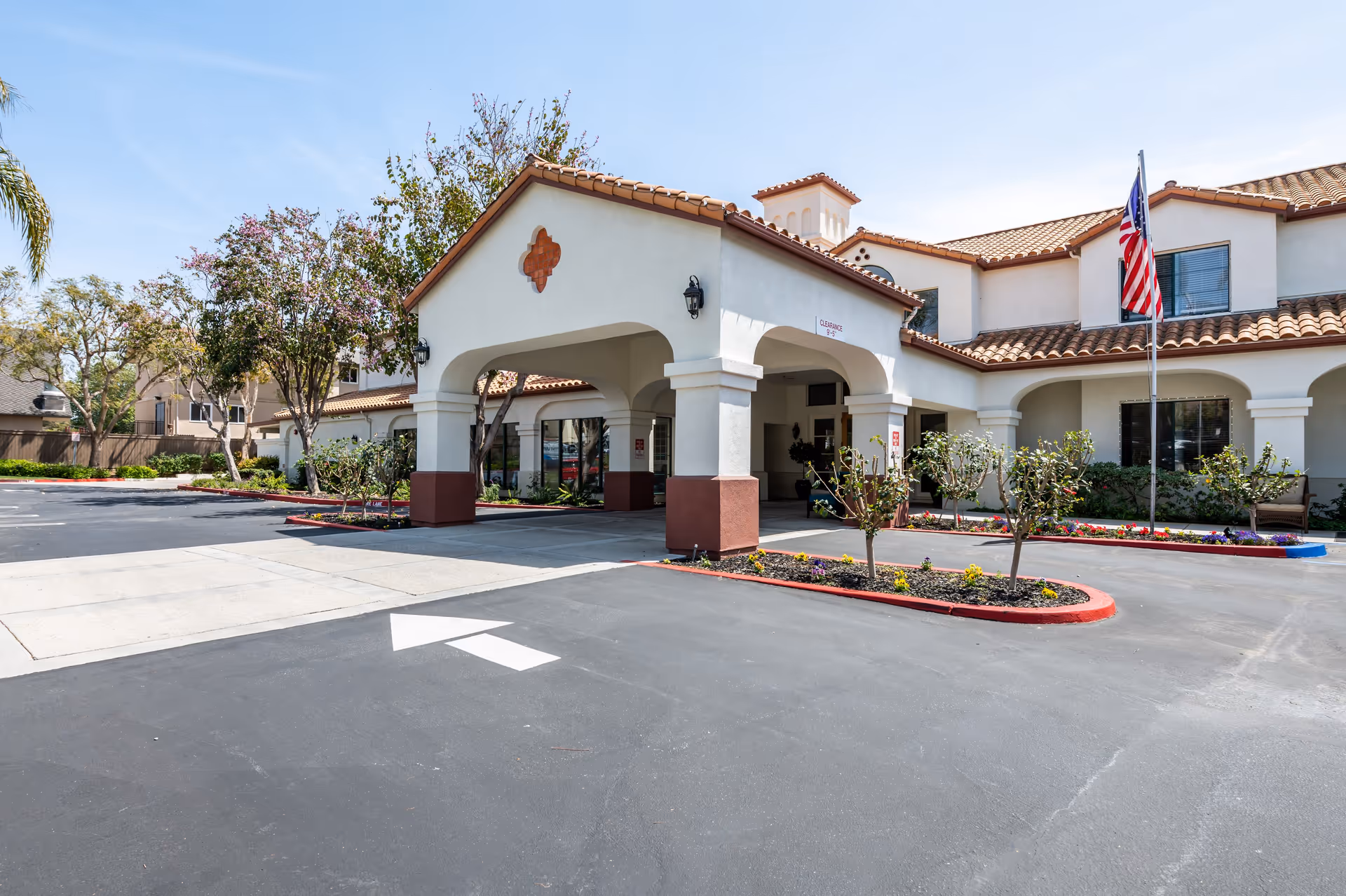 Exterior view of Aegis Living Ventura facility showing the entrance with a covered driveway, landscaped flower beds, an American flag on a flagpole, and a clear blue sky.