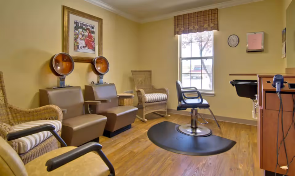 Salon-style interior with two hooded hair dryers, a styling chair on a floor mat, wicker chairs, and a window.