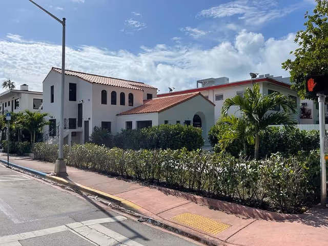 Exterior view of a two-story building with white walls and red tile roofs, surrounded by green bushes and palm trees under a partly cloudy sky. A sidewalk and street with a traffic light are visible in the foreground.
