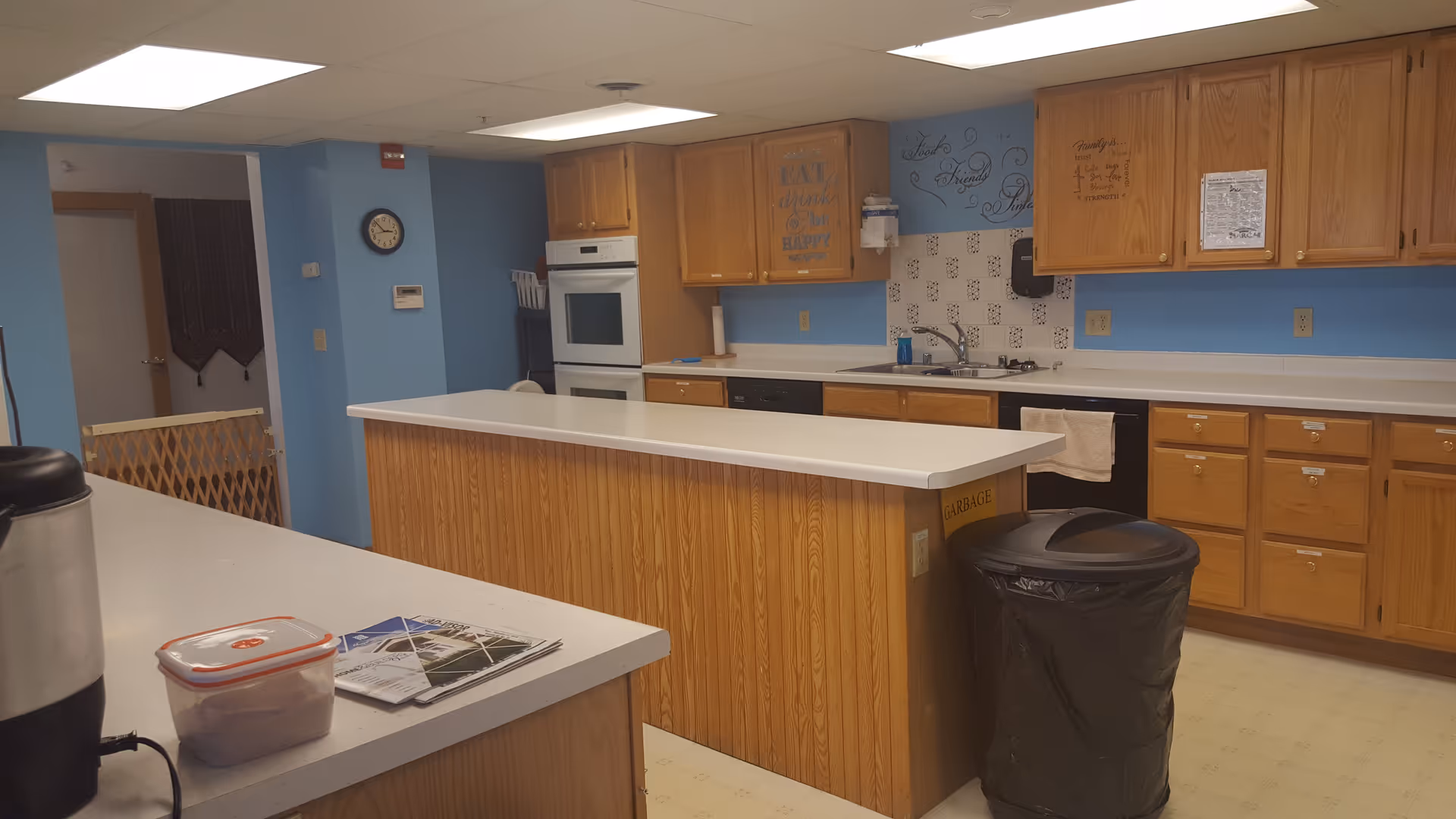 Communal kitchen with a long center island, wood cabinetry, blue walls, an oven and sink, and a trash can.