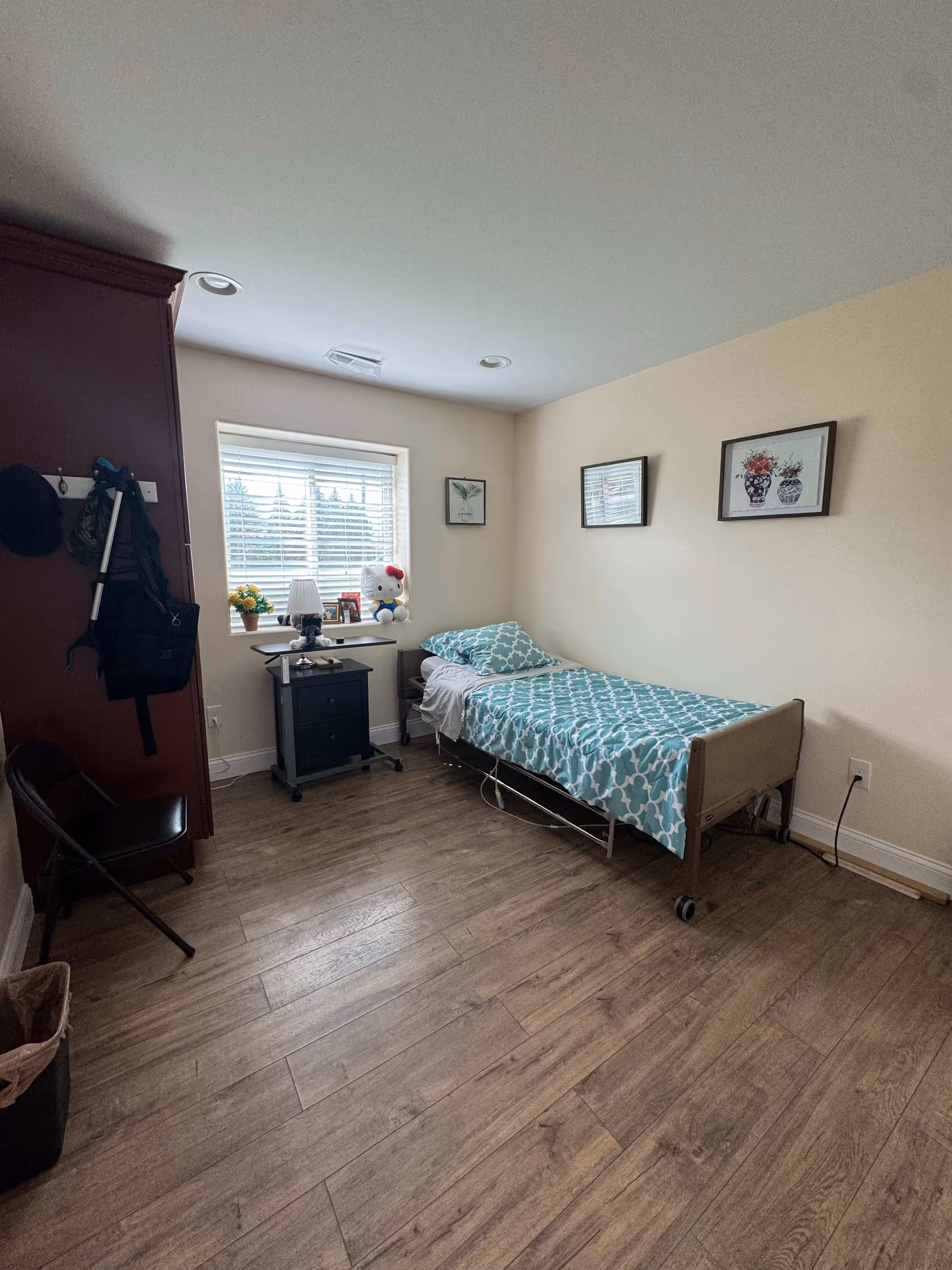 A small bedroom with a single bed covered in a blue and white patterned bedspread. Next to the bed is a black nightstand with a lamp, a small plant, and a Hello Kitty plush toy on the windowsill. The room has wooden flooring, beige walls with three framed pictures, and a window with white blinds letting in natural light. There is a dark wooden wardrobe on the left side with some items hanging on hooks and a folding chair nearby.