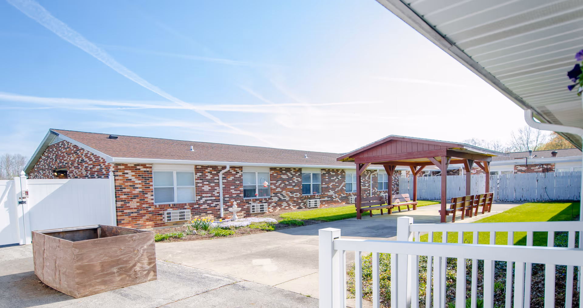 Outdoor courtyard area of a senior living facility with a brick building in the background, a wooden gazebo with benches on a concrete patio, green grass, and a white fence surrounding the area under a clear blue sky.