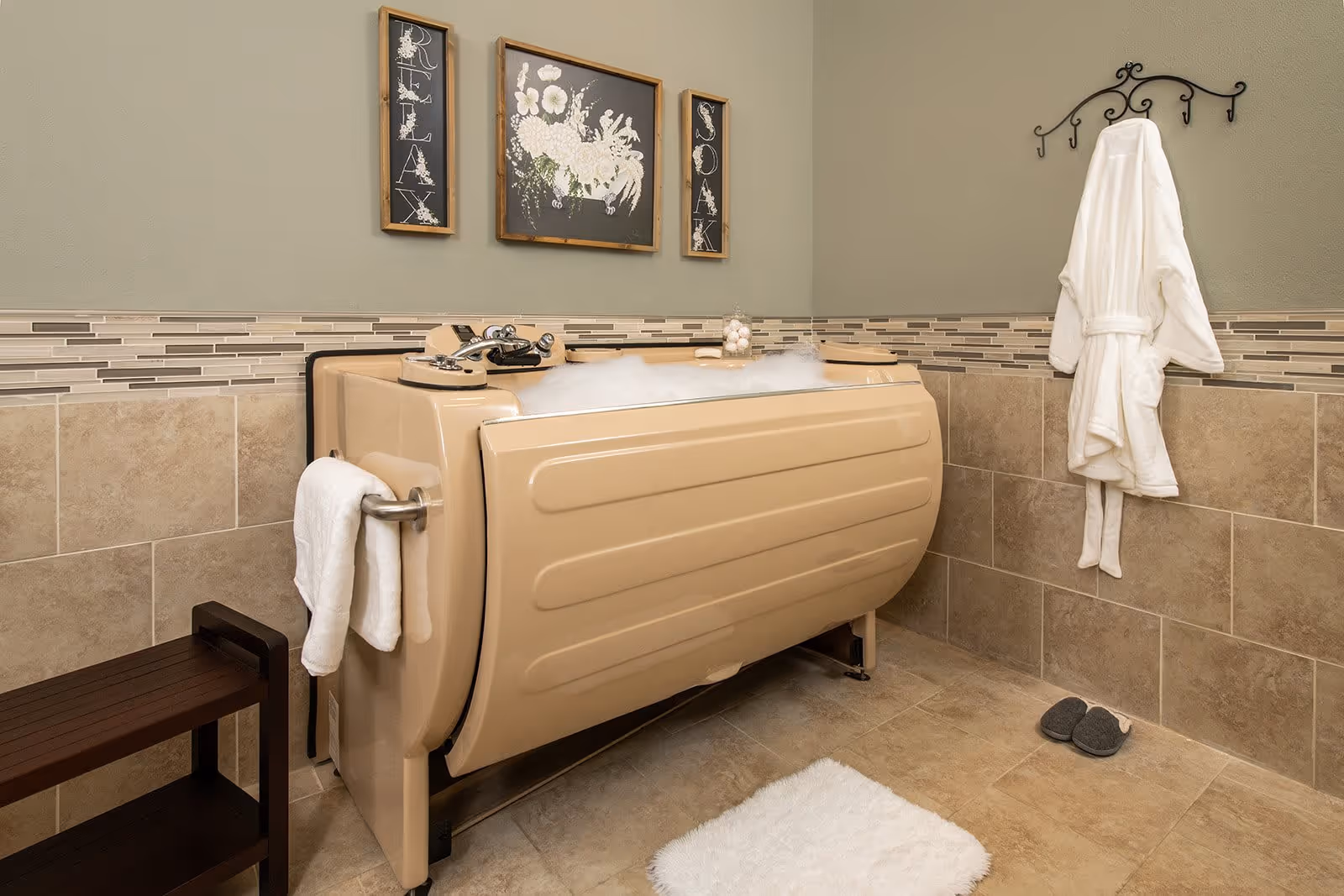 A beige walk-in bathtub filled with bubbles in a tiled bathroom. A white towel hangs on a bar attached to the tub, and a white bathrobe hangs on a decorative black wall hook. There are three framed wall decorations above the tub, with the words 'RELAX' and 'SOAK' on the side frames and a floral image in the center. A small dark wooden bench is next to the tub, and a white bath mat and gray slippers are on the floor.