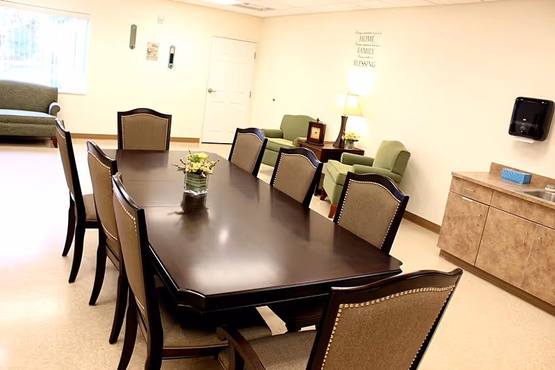 A well-lit room with a long dark wooden dining table surrounded by eight upholstered chairs. In the background, there are two green armchairs, a small side table with a lamp, and a wall decal with an inspirational quote. To the right, there is a countertop with cabinets, a sink, and a wall-mounted paper towel dispenser. A window with vertical blinds is visible on the left side of the room.