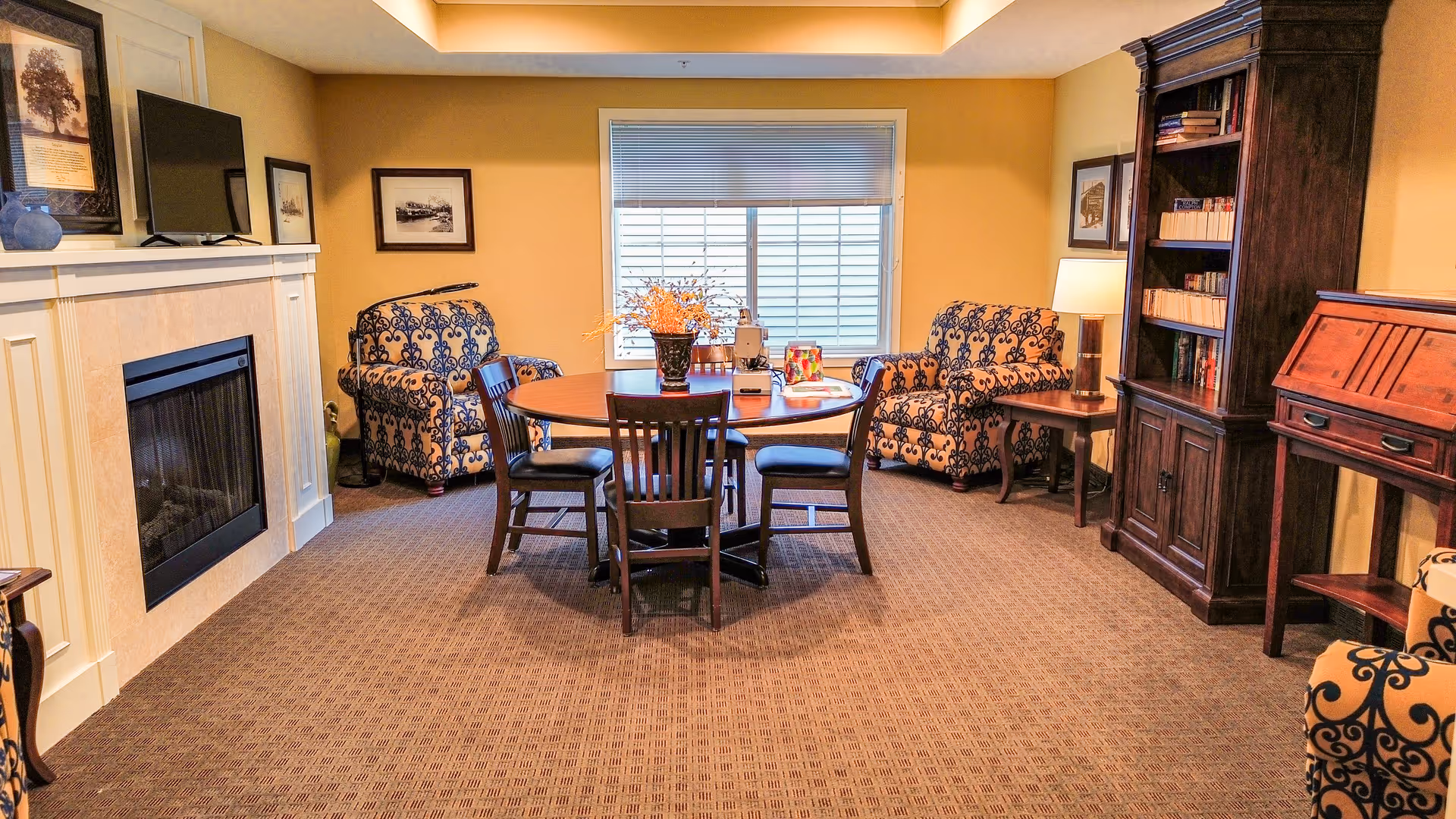 Cozy common room with a round table and chairs, patterned armchairs, a fireplace, and a bookcase under a window.