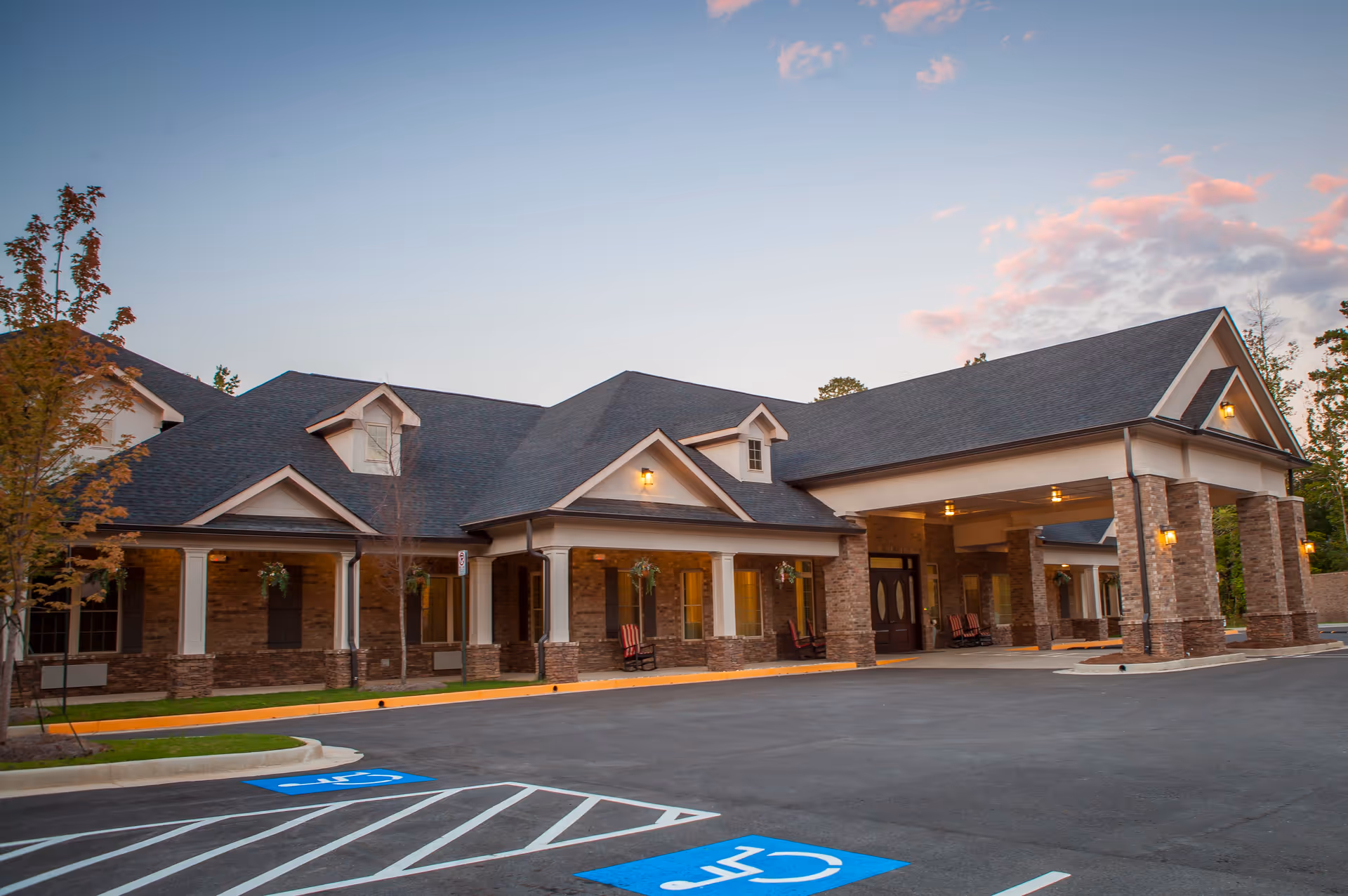 Front exterior of a single-story brick senior living facility with a covered porte-cochère entrance and visible handicap parking spaces at dusk.