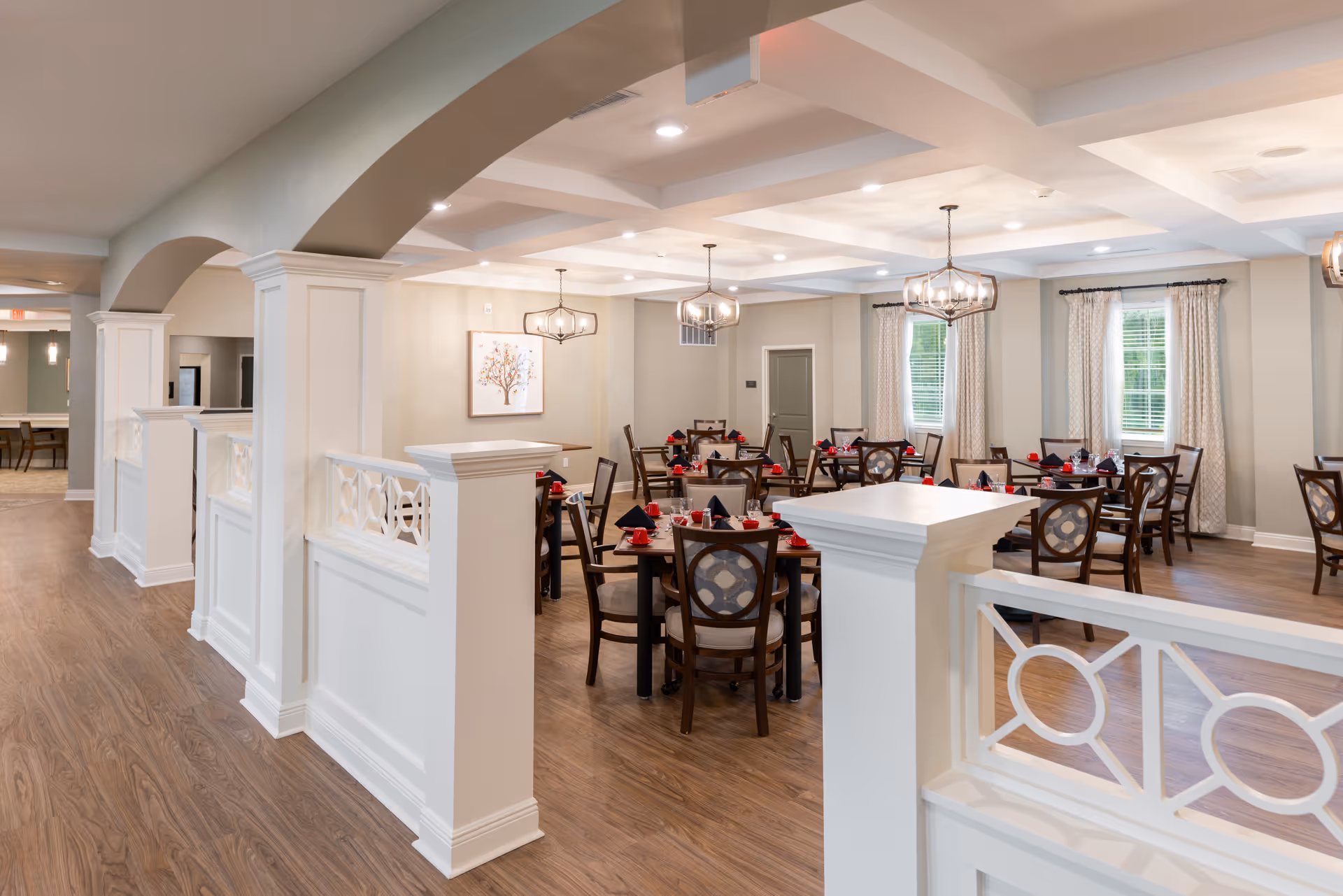A spacious dining room with multiple round tables set with red napkins and black tableware. The room features wooden floors, white walls with decorative molding, large windows with curtains, and modern pendant lighting fixtures hanging from a coffered ceiling. White half-walls with decorative cutouts and arches separate the dining area from the adjacent hallway.