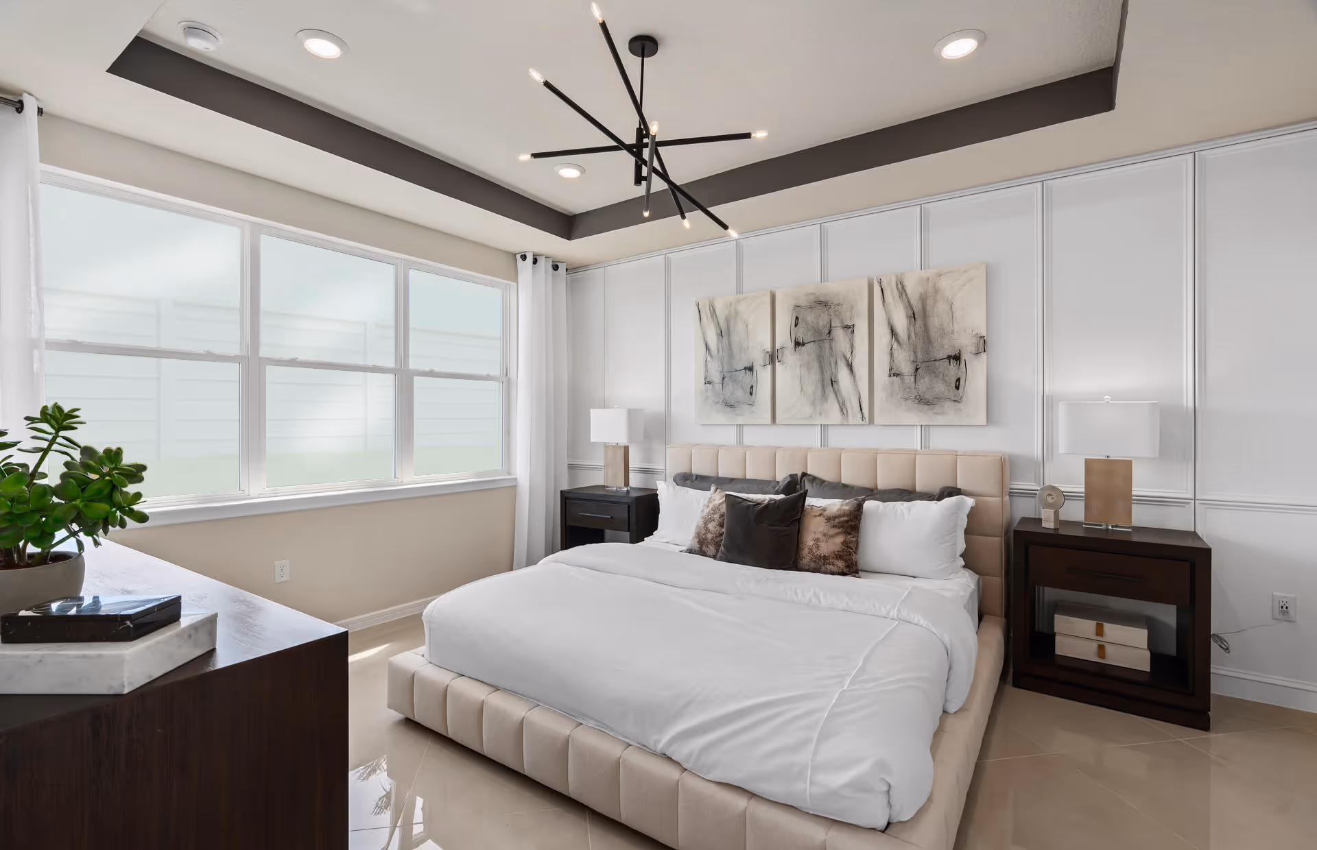 A modern bedroom with a large beige upholstered bed featuring white and brown pillows. The room has a dark wood dresser with a plant and decorative items on top, two matching dark wood nightstands with lamps, and abstract artwork on the white paneled wall behind the bed. A large window with white curtains lets in natural light, and the ceiling has recessed lighting and a contemporary black chandelier.