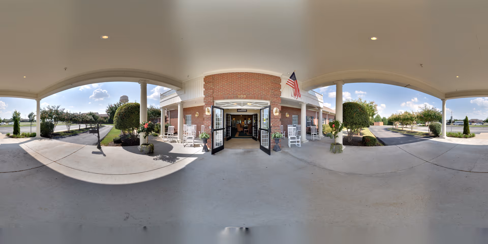 Covered front entrance and drive-up of a brick senior living building with rocking chairs, potted plants, an American flag, and open double doors.